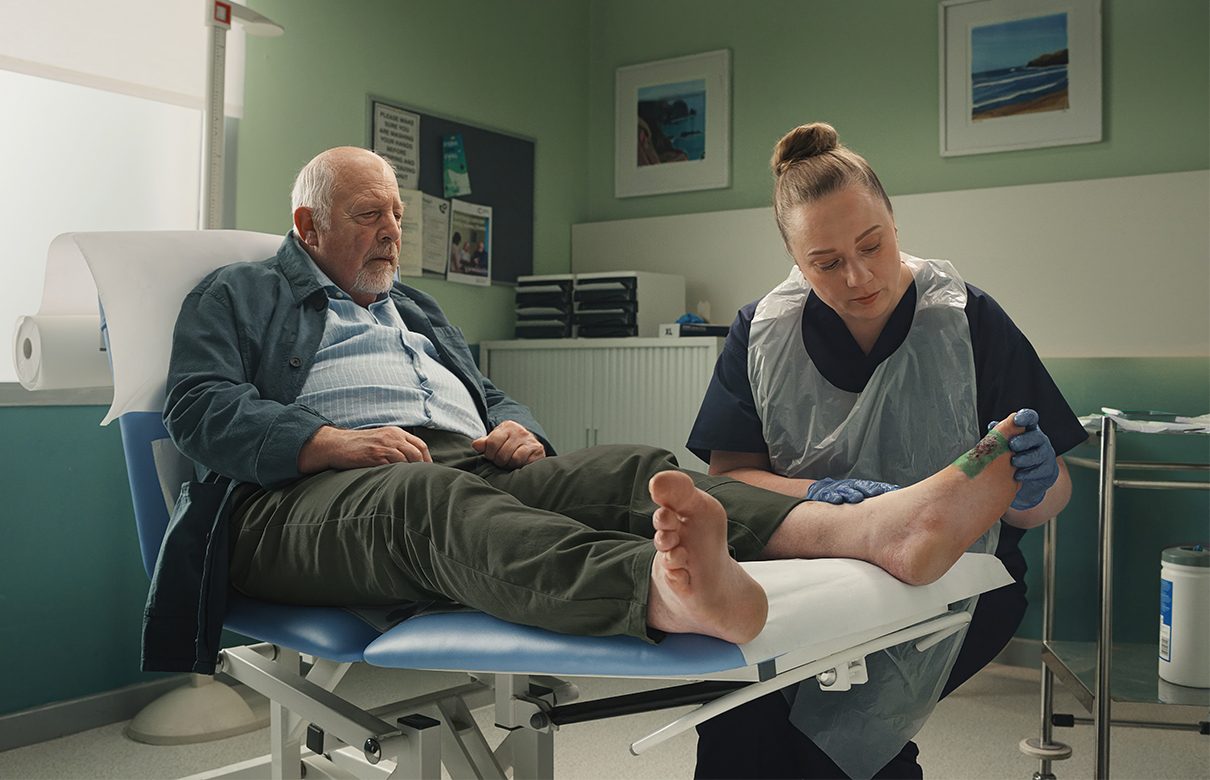 Helsepersonell i et behandlingsmiljø legger på en Sorbact-bandasje på foten til en eldre mann.